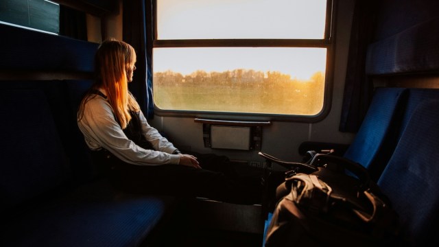 Solo travel for women and members of the LGBTQ community represented by a woman sitting alone in the car of a train, staring out the window on the bright fields passing by.