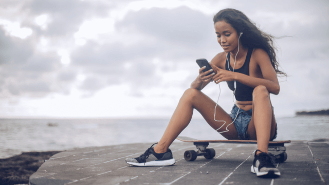 A young woman sitting on a skateboard and listening to Indonesian podcasts with her phone
