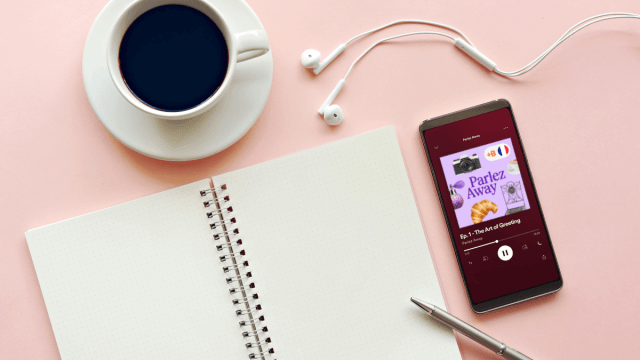French podcasts represented by an iPhone playing a Parlez Away episode along with a cup of coffee and a notebook with a pen nearby.