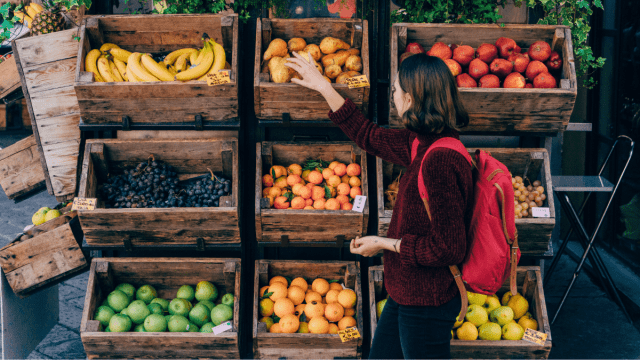 Food idioms represented by a woman at a fruit market standing in front of an array of wooden boxes holding limes, lemons, bananas and other fruits.