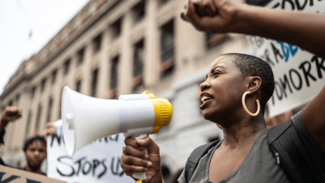 Woman with megaphone standing in front of a government building and a few protest signs that can't be fully read.