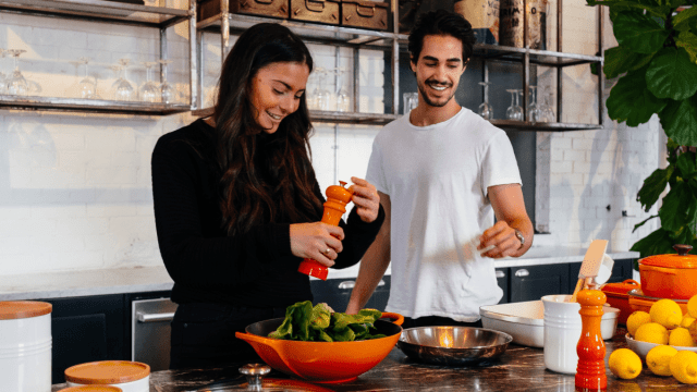 Cooking and language learning represented by a young man and woman in their kitchen preparing a meal with lettuce.
