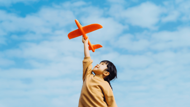 Learn a language like a child represented by a young child in a sweater holding up a plastic orange airplane against a partly cloudy sky.